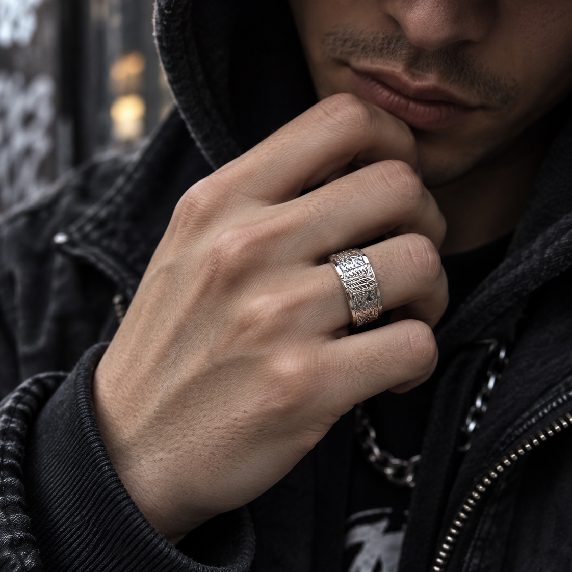 A close-up lifestyle shot of a man wearing a wide silver band ring with a textured, patterned surface. He is wearing a black zip-up jacket over a black hoodie, with his hand resting near his chin, showcasing the ring.
