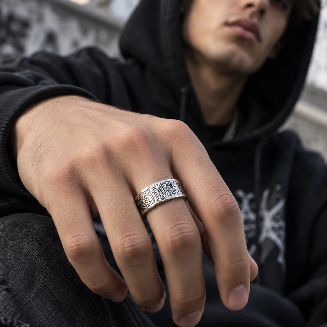 A close-up of a man wearing a detailed silver ring on his ring finger, dressed in a black hoodie and dark jeans, sitting outdoors.