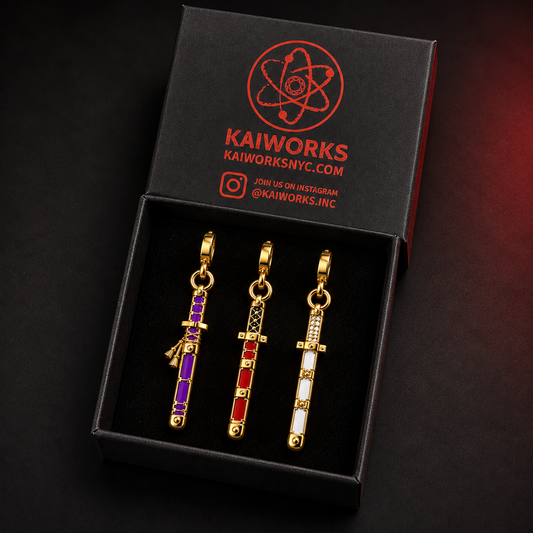 Three gold-tone sword-shaped jewelry charms with purple, red, and white enamel details displayed in a black KAIWORKS gift box.