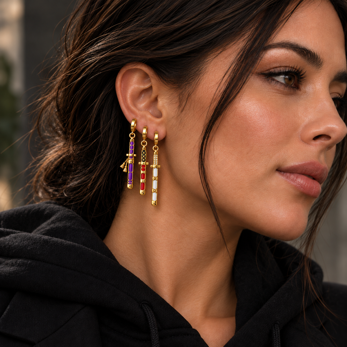 A close-up of a woman wearing gold drop earrings with red, purple, and white crystal accents, paired with a black hoodie.