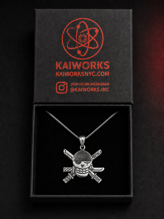 A silver skull and crossbones pendant necklace displayed inside an open black gift box with a branded black lid. The lid features red printed branding for KAIWORKS and social media details.