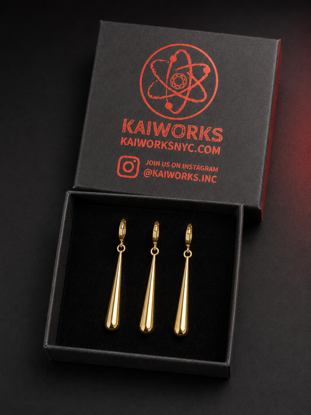 Three gold teardrop-shaped earrings displayed in an open black presentation box with a branded lid. The lid shows the brand name KAIWORKS and social media text in red.