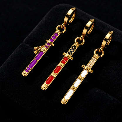 Three gold-tone sword-shaped earrings displayed on a black velvet jewelry box. Each earring features a gold clasp and a vertical sword design with rectangular colored inlays: one purple, one red, and one white.