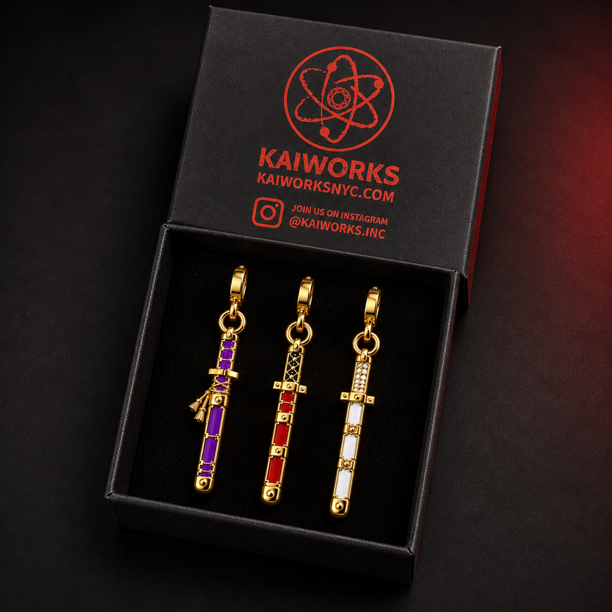 Three gold-tone sword-shaped jewelry charms with purple, red, and white enamel details displayed in a black KAIWORKS gift box.