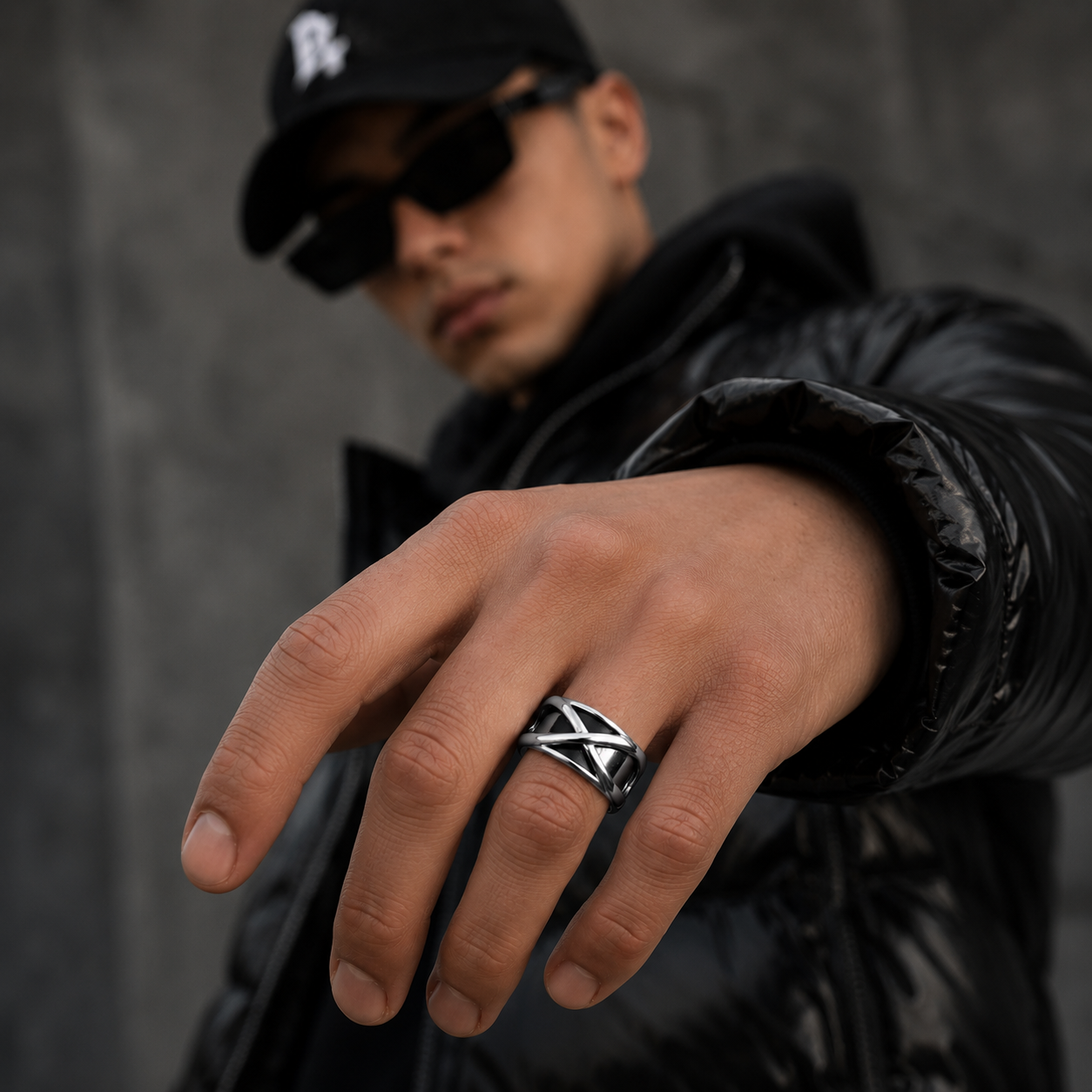 Man wearing a black cap and sunglasses, showing a silver ring on his finger against a gray background.