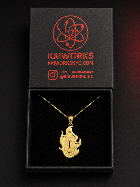 A gold pendant necklace with a flame-like design is displayed inside an open black presentation box. The box lid features the red KaiWorks logo and branding.