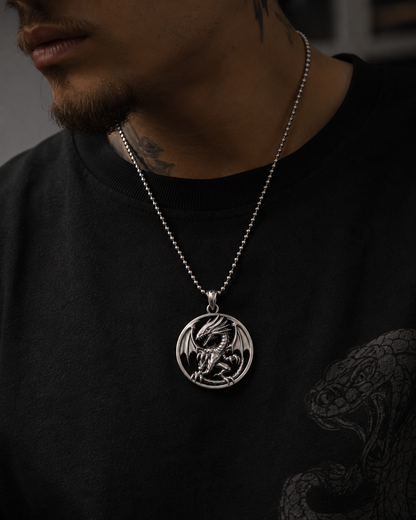 A close-up of a man wearing a silver dragon pendant necklace on a black beaded chain, styled with a black crewneck shirt featuring a snake graphic.